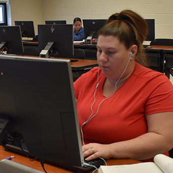 Female Student in computer lab wearing headphones with student on computer in background